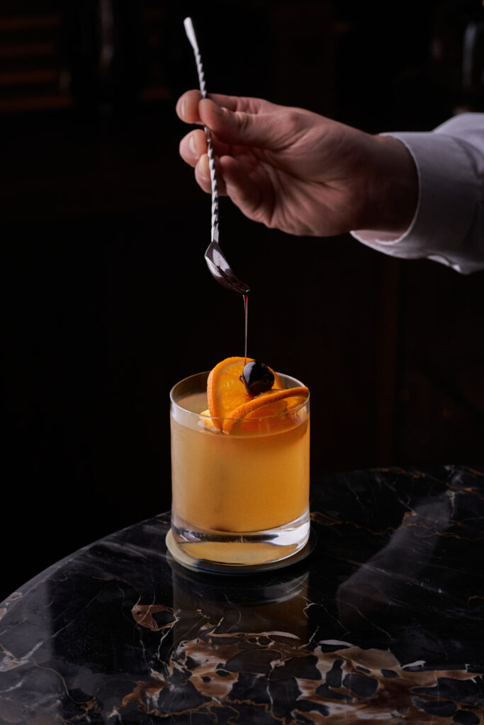 Image of a hand holding a silver stirring spoon above an orange punch cocktail garnished with orange slices